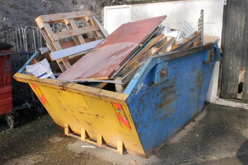 A large open-top metal skip painted blue with visible rust and weathering, situated outdoors on a paved surface adjacent to a white wall and black wooden fencing. The skip is filled with an assortment of discarded waste materials including wooden pallets, metal sheets, and other construction debris, some of which are leaning or stacked precariously. The wooden pallets are light brown with rough textures, while the metal sheets exhibit a reddish-brown corrosion finish. The debris shows varying textures, from smooth painted surfaces to rough wooden textures. In the background, there is a grassy area and a pile of additional waste on the left side. The scene appears to be part of a rubbish removal operation, typical of garden clearance or waste disposal services offered by Garden Clearance Willesden in the NW London area, near Willesden or surrounding postcodes.