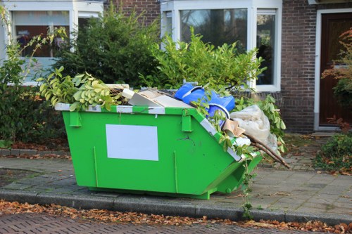 A large, bright green waste skip positioned on a paved driveway in front of a residential building with brick walls and white-framed windows. The skip is filled with a mixture of green foliage, tree branches, and various discarded materials, including cardboard boxes, white plastic packaging, and some bagged rubbish. Several leaves and small branches spill over the top edge of the skip, which appears to be used for garden clearance. In the background, shrubbery and small trees are visible, with some greenery extending above the skip. The surrounding pavement is clean, with a curb separating it from the street, and the scene suggests a typical suburban environment. The image aligns with rubbish removal services such as those provided by Garden Clearance Willesden, highlighting the collection of garden waste and general rubbish from a residential property in the area.