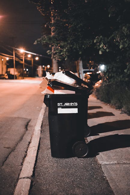 A black wheeled rubbish bin labeled 'ST. JOHN'S' positioned on the edge of a paved sidewalk beside a residential street at night. The bin is filled with various discarded items, including cardboard bo