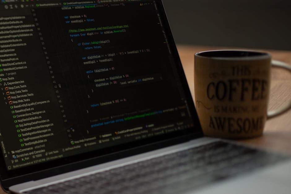 A close-up photograph of a laptop computer displaying lines of source code on a dark background, positioned on a wooden desk surface. To the right of the laptop, there is a ceramic coffee mug with a b